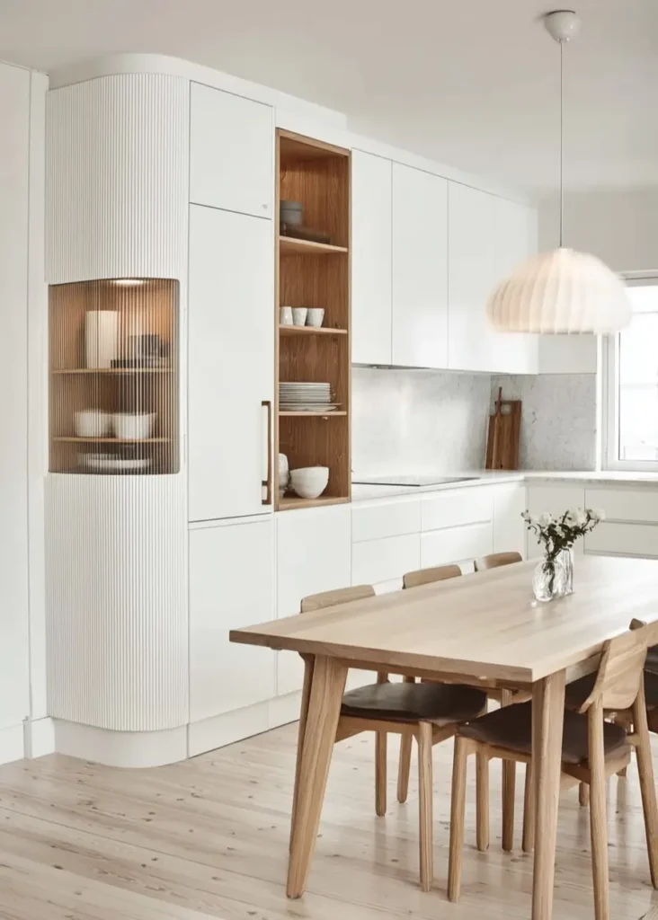 A contemporary Scandinavian kitchen with white cabinets, a curved fluted-wood display niche, and a light wood dining table.