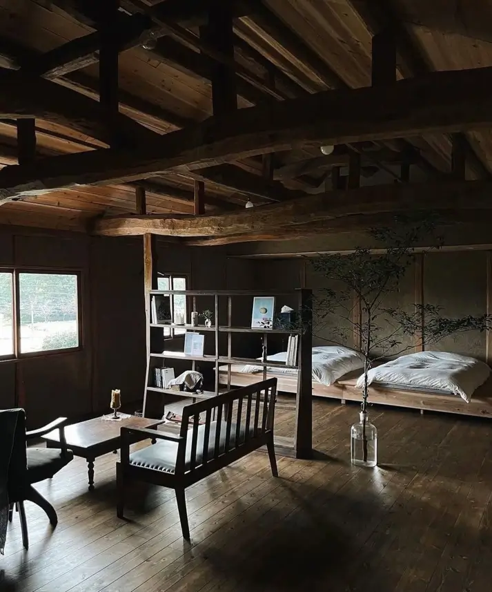 A rustic Japanese farmhouse (kominka) interior with dark exposed beams, wooden floors, and simple furniture.