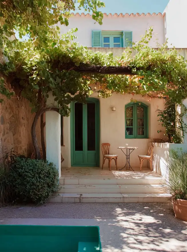 38 Small Mediterranean-Style Homes That Define Coastal Charm 31 Cream stucco courtyard with a leafy grapevine canopy, emerald green French doors, and simple patio seating.