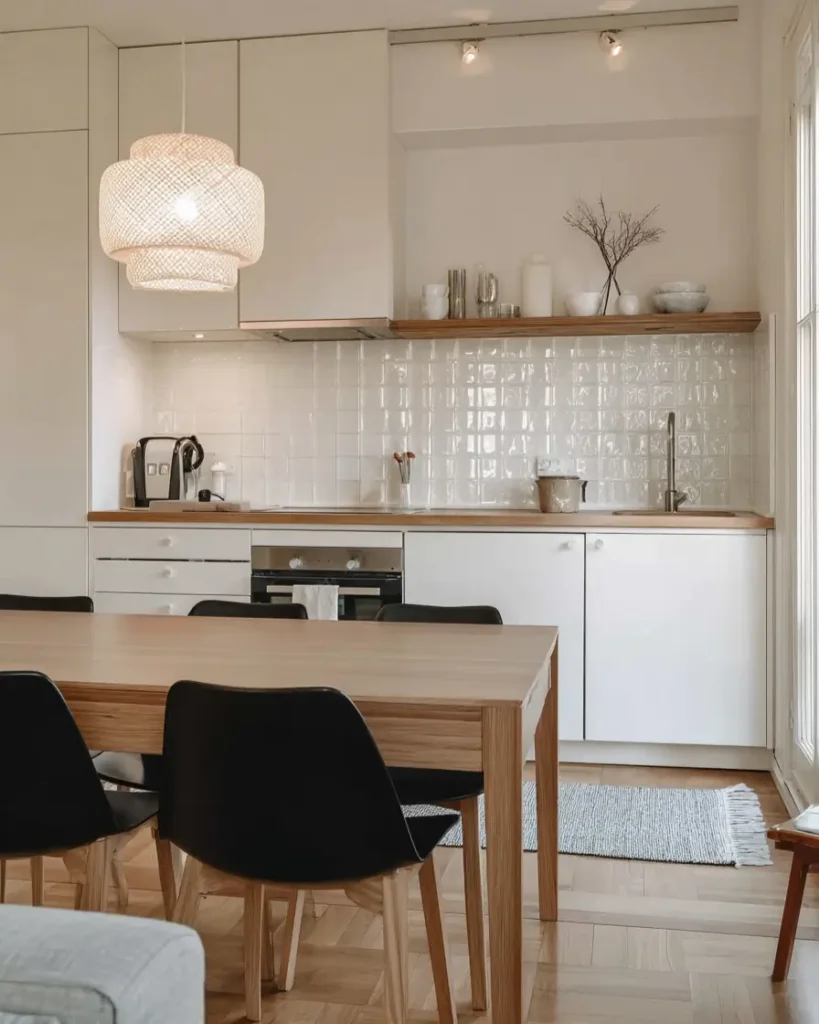 A contemporary Scandinavian kitchen featuring white cabinets, a glossy white tiled backsplash, and a wood dining table with black chairs.