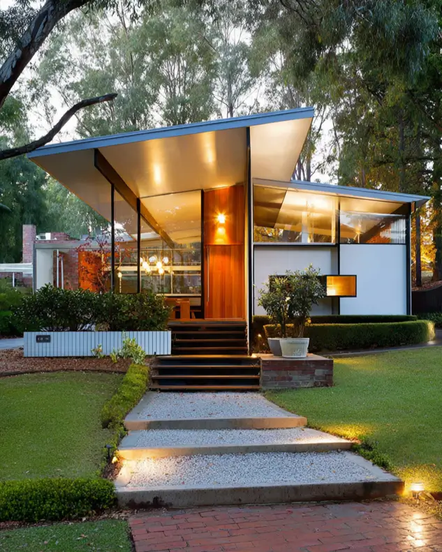 37 Mid Century Modern House Exteriors to Inspire Your Renovation 33 A mid century modern house exterior at dusk with a sculptural gull-wing roof, illuminated glass walls, and a wood door.