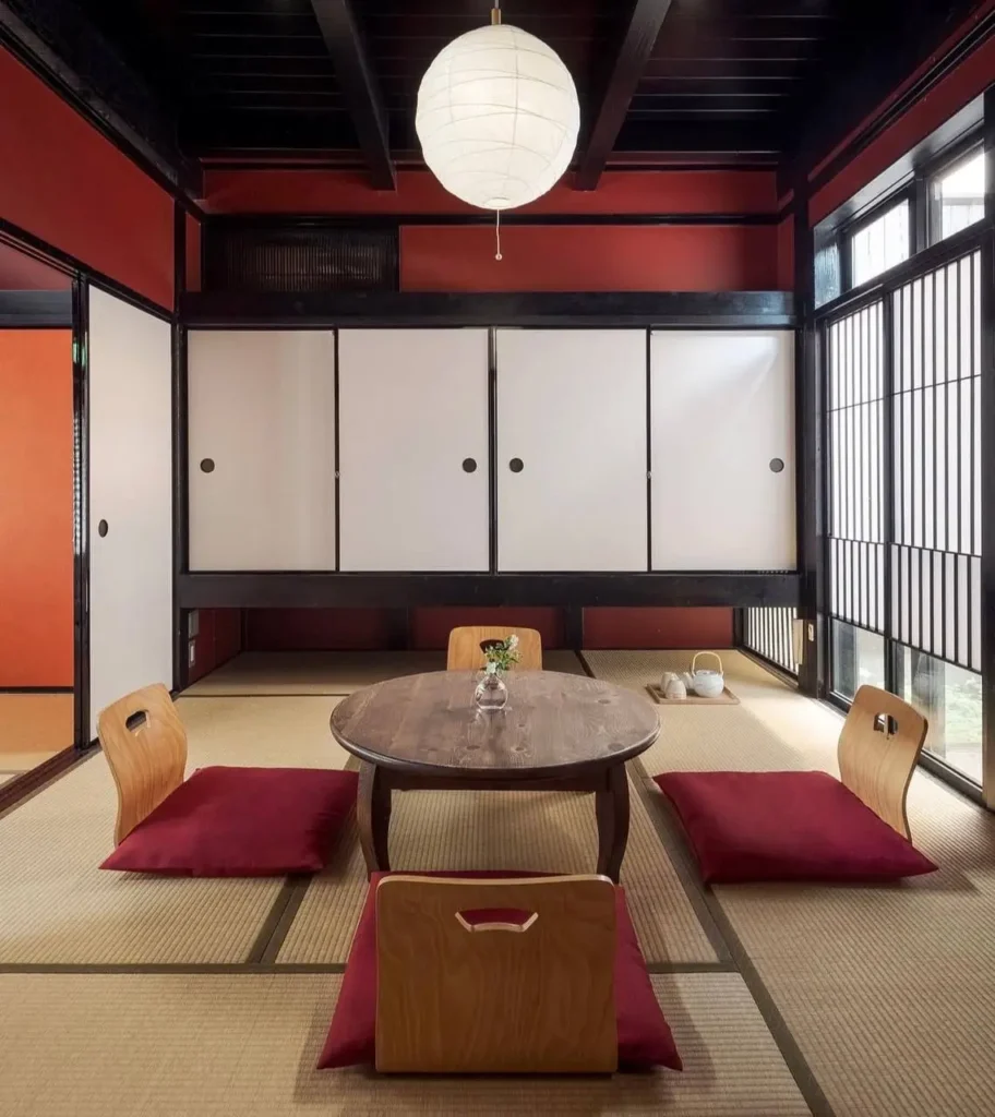 A traditional Japanese tatami room featuring historic 'suo' red walls, a dark beam ceiling, and red floor cushions.