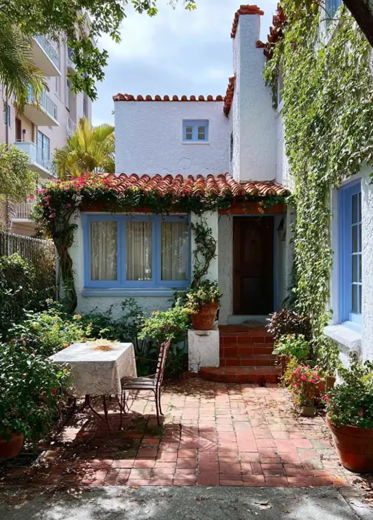 38 Small Mediterranean-Style Homes That Define Coastal Charm 39 White stucco small Mediterranean house design featuring a hidden brick patio, intimate seating area, and lush green privacy screen.