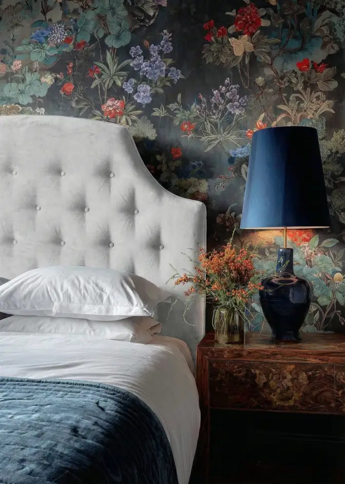 49 Headboard Design Ideas That Instantly Elevate Your Bedroom 48 Light grey button-tufted headboard against dark moody floral wallpaper with blue lamp.