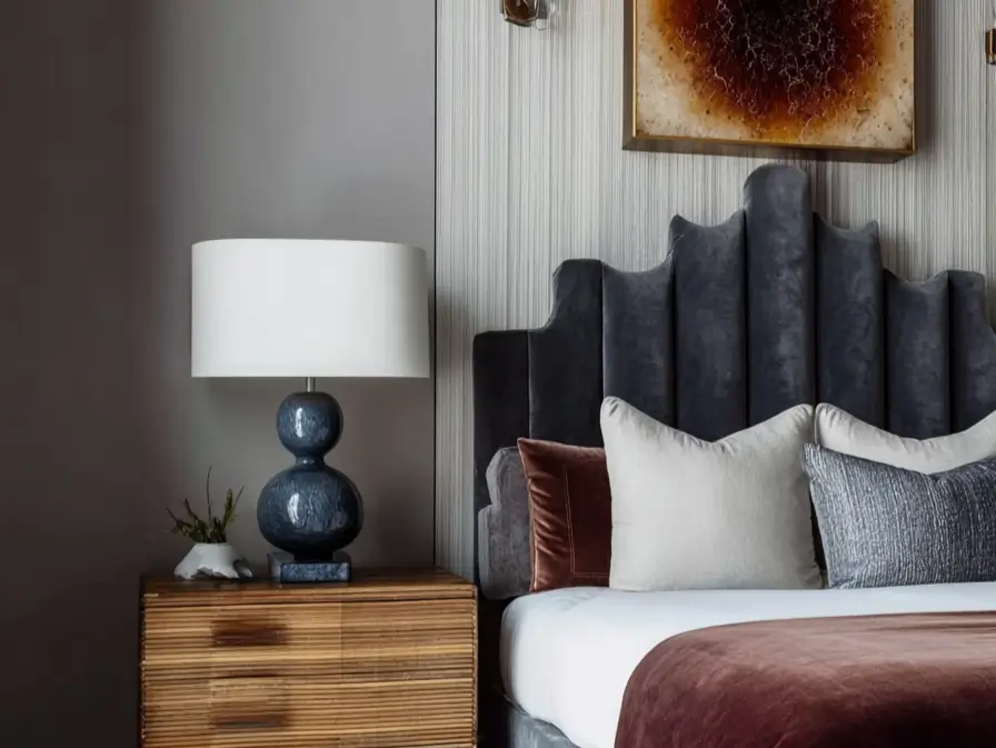 Charcoal grey velvet headboard with Art Deco stepped silhouette and channel tufting, paired with a sculptural blue lamp and ribbed wooden nightstand.