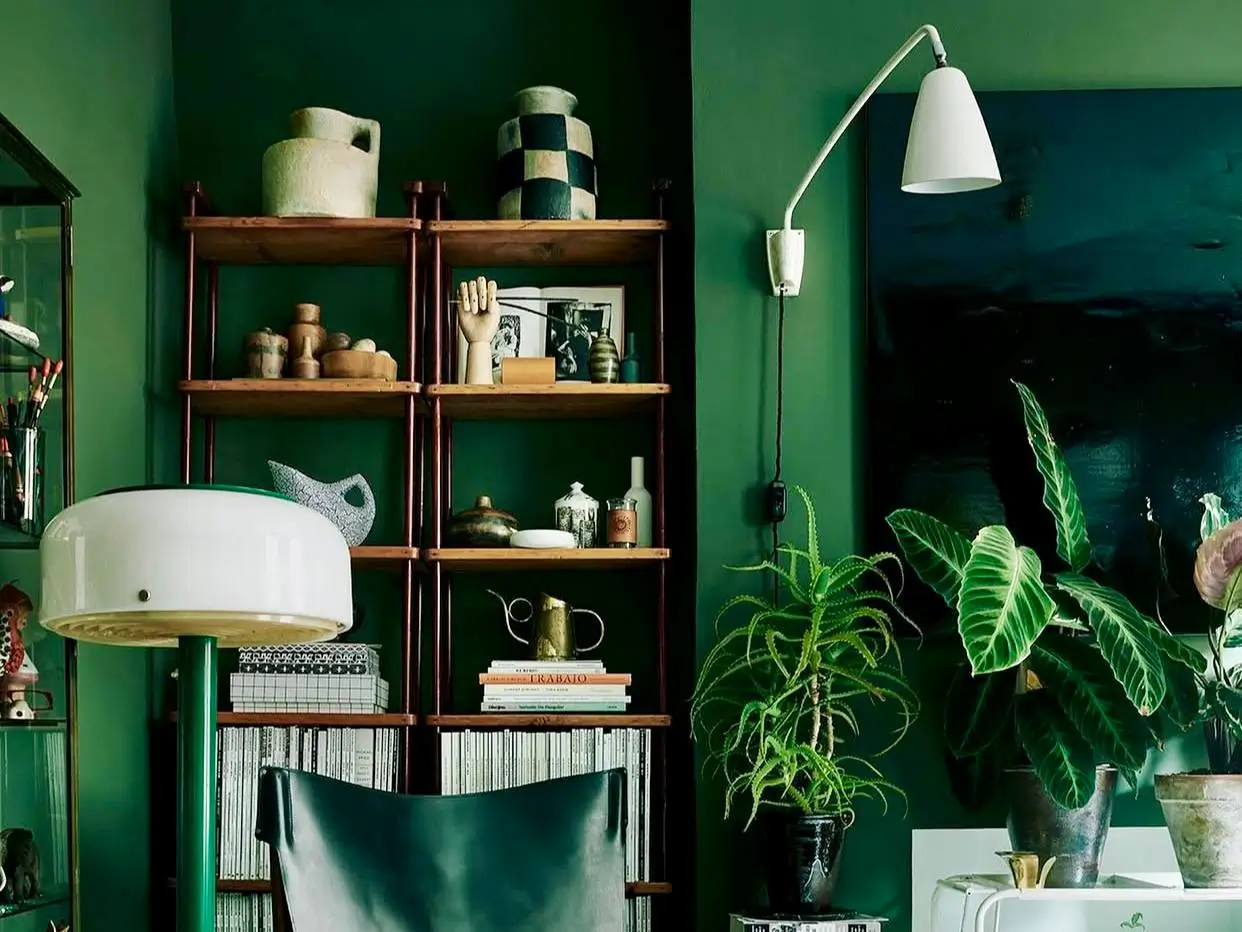 A sophisticated study with deep green interior house paint colors on the walls, a wood bookshelf with pottery, and a modern white wall lamp.