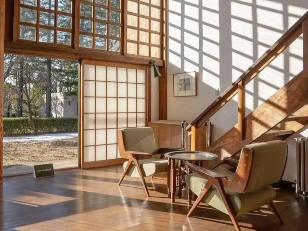 A sunlit Japandi style home interior with large shoji-style grid windows, warm wood floors, and two mid-century modern chairs.
