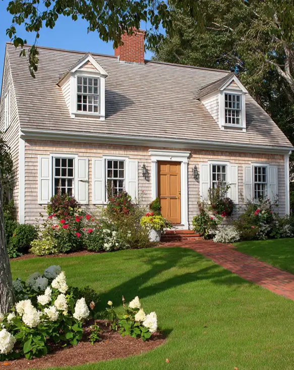 27 Cape Cod Style House Ideas for a Timeless New England Exterior 21 Cedar shake cottage with cream shutters, two pedimented dormers, mustard-yellow front door, rose garden beds, and herringbone brick path