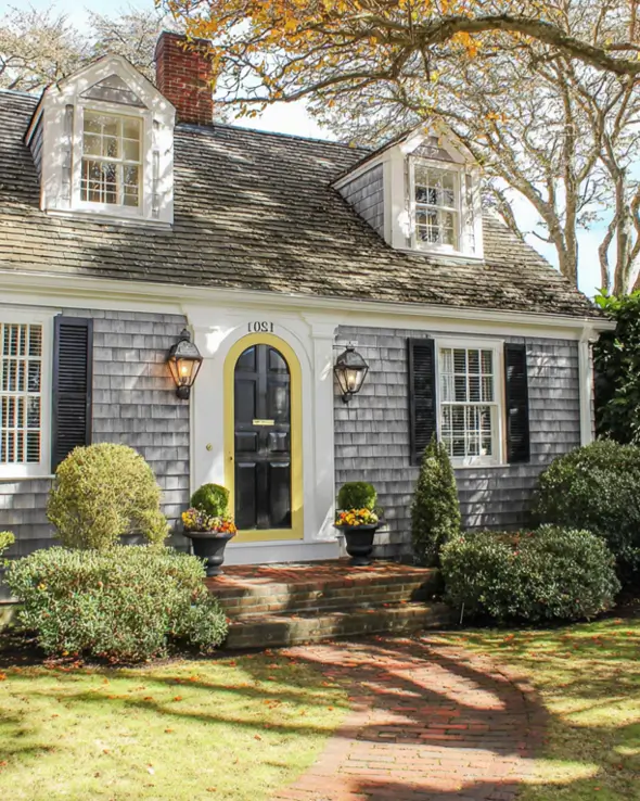 27 Cape Cod Style House Ideas for a Timeless New England Exterior 25 Gray shake cottage with two pedimented dormers, black shutters, arched black front door framed in yellow, brick steps, and autumn urns