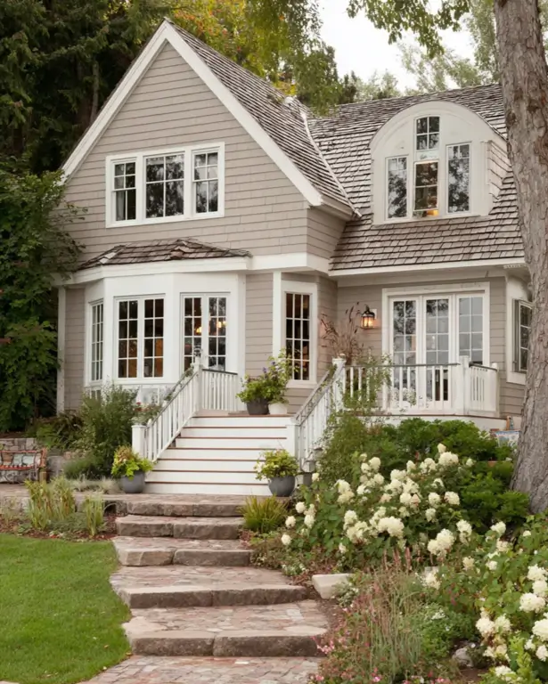 27 Cape Cod Style House Ideas for a Timeless New England Exterior 26 Greige shingled cottage with layered gables, curved barrel dormer, bay windows, white deck railings, and stone garden steps