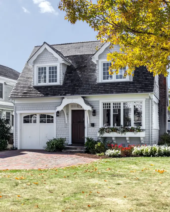 27 Cape Cod Style House Ideas for a Timeless New England Exterior 9 White shingled cottage with carved bracket entry hood, arched garage door, mauve front door, and red flower window box