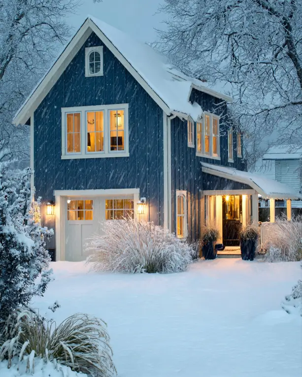 33 Blue and White Cottage Ideas That Always Feel Fresh 17 navy blue and white cottage exterior with navy board-and-batten siding, white trim, lit windows, and falling snow at dusk