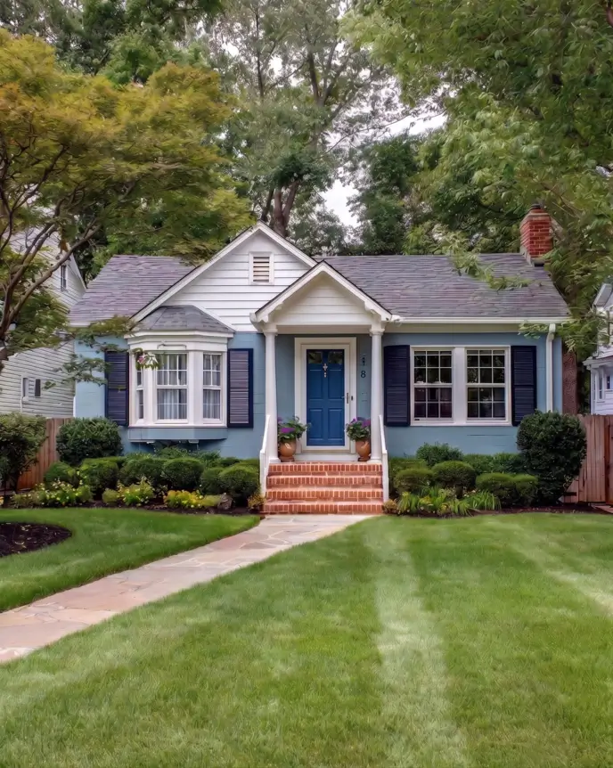 33 Blue and White Cottage Ideas That Always Feel Fresh 11 blue and white cottage exterior with dusty blue-grey walls, navy shutters, white bay window, and a columned brick-step entry