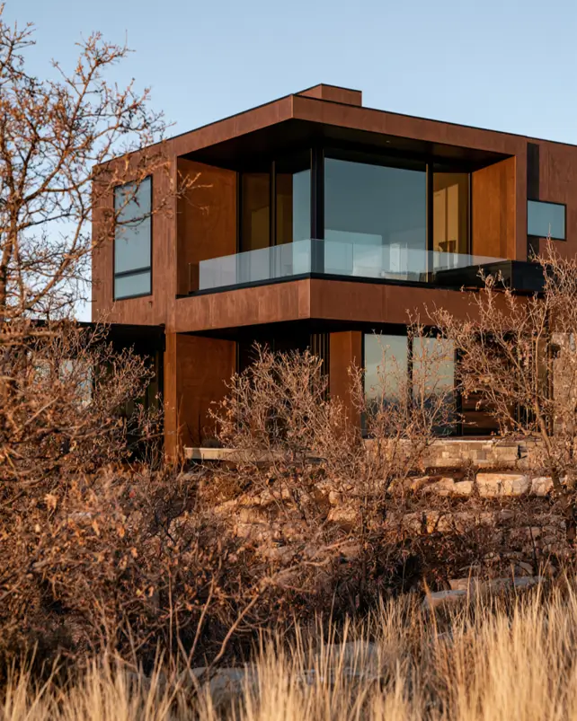 37 Brown House with Black Trim Ideas for a Warm, Modern Exterior 5 Weathered Corten steel home with a wraparound charcoal-framed corner window on a stone base at dusk