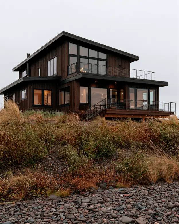 37 Brown House with Black Trim Ideas for a Warm, Modern Exterior 38 Deep chocolate-brown stained wood coastal home with black window frames and charcoal deck railings above a cobble beach