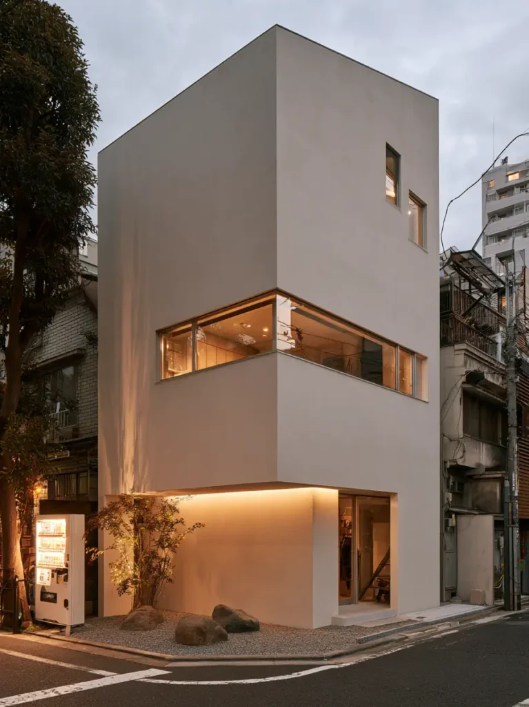 Modern Japanese House Design: 58 Ideas, Costs, and What Defines the Style 8 A lit window wrapping the corner of a pale render house, with placed rocks and a slim tree.